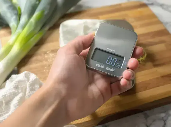 A hand placing a leek on a digital scale, symbolizing the monitoring of food intake for weight management.